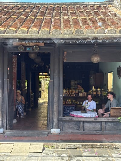 This image depicts the entrance to a teahouse in Hội An Ward, Vietnam. The teahouse has a traditional tiled roof and wooden facade. Two men are seated at a table inside the teahouse, one holding a teacup and the other looking out. A woman is seated to the left of the entrance. Shelves stocked with jars of tea are visible in the background. The street in front of the teahouse is paved with stone.