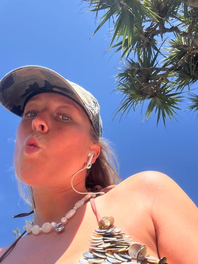 A low-angle, close-up photograph features a young female posing for the camera under a clear, bright blue sky, indicative of a sunny day. Her face, seen from slightly below, is angled towards the viewer, and she sports a playful "pouty" expression with her lips puckered. Her light-colored eyes are open and directed at the camera.
She wears a camouflage-patterned bucket hat covering the top of her head and white wired earbuds in both ears. Around her neck is a necklace composed of various white, possibly pearlescent, beads and silver-toned accents. On her right side, a portion of her shoulder and upper arm is visible, along with what appears to be a top or swimsuit strap embellished with small, shell-like decorative elements.
The background primarily consists of a vibrant blue sky, with the lush green foliage of a tropical plant, possibly a palm or pandanus tree, visible in the upper left quadrant. The bright lighting casts a warm glow on her skin. The scene suggests a casual, outdoor setting in a warm climate, consistent with the specified location of Coolangatta, Australia. There is no visible text in the image.