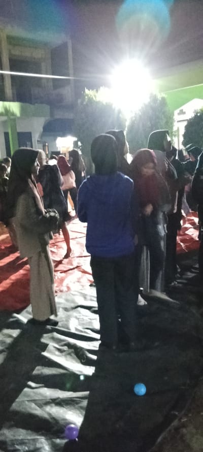 The image shows a group of people standing on a black tarp in front of a building at night. The people are standing in a dimly lit area with a large light source shining brightly in the background. The light source creates a glare that obscures some of the details of the people and the surrounding environment. There are eight people in the image. The people are dressed in dark clothing with the exception of one person wearing a light blue jacket and another wearing a red scarf. The people appear to be talking and interacting with each other. There are two small, colorful balls on the ground in the foreground of the image, one blue and one purple. The image was taken in Cangkuang, Indonesia, at coordinates -7.065 latitude, 107.552 longitude. The image is taken from a low angle, looking up at the people. The background is blurry due to the light source. The image is slightly out of focus, but the overall scene is clear.