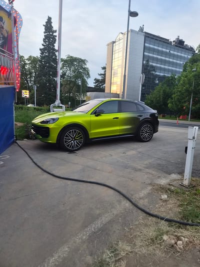 The image captures a green Porsche Cayenne parked on the side of a road in Esch-sur-Alzette, Luxembourg. A large modern building and trees are visible in the background. A black cable runs along the edge of the pavement near the car. In the foreground, a portion of a fairground ride with blue and red lights is visible.