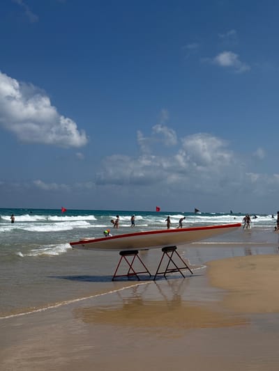 A beach scene in Bat Yam, Israel, under a bright blue sky with scattered white clouds. The ocean is visible with waves rolling onto the sandy shore. In the foreground, a long, narrow boat, possibly a surf ski or a rescue boat, is resting on a stand on the sand. It is predominantly white with a red trim and appears to be made of fiberglass. Several small red flags are planted in the sand near the water's edge.
There are a number of people visible on the beach and in the shallow water, some appearing to be swimmers or beachgoers. A few individuals are in or near the water, and others are on the sand. One person is wearing a bright green helmet. The presence of flags and the boat suggests a possible water sports event, training session, or beach patrol activity.
The lighting indicates it is daytime, likely late morning or afternoon, given the shadows cast by the boat's stand. The weather appears to be sunny and pleasant, typical of a beach day. The overall atmosphere seems active and recreational. No visible text is present in the image.