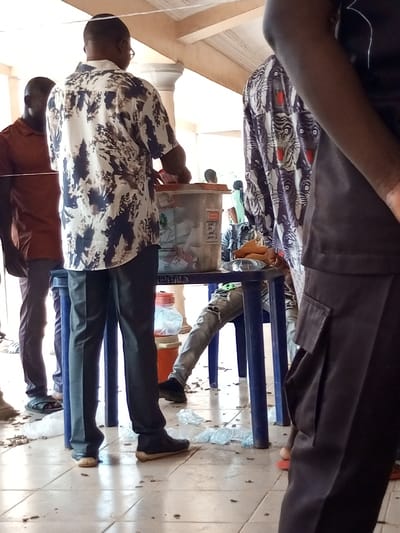 The scene depicts an interior or semi-enclosed exterior area in Ajalli, Nigeria, characterized by a tiled floor and structural columns. The floor is covered with scattered debris, predominantly empty clear plastic water bottles and dark organic matter.
In the central foreground, a dark-skinned male, observed from the rear, stands upright. He wears a short-sleeved shirt featuring a white and dark-toned abstract pattern, and dark trousers with dark footwear. His right hand is positioned over the opening of a translucent plastic container with an orange lid, which rests on a blue plastic table. The container appears to hold assorted papers or similar items, and has a white label with red text attached to its side.
To the immediate left of the central figure, a second dark-skinned male stands partially visible, wearing a reddish-brown short-sleeved polo shirt and dark trousers. A red bucket is situated on the floor near the central figure's left foot.
To the right of the central figure, partially obscured by an individual in the immediate foreground (only a dark arm and leg with a dark, possibly pleated, garment and red footwear are visible), a third person is seated on a blue plastic stool. This individual wears light-colored distressed jeans and dark shoes; only their lower torso and legs are distinctly visible. On the blue table near this seated person, indistinct light-colored objects are present.
In the mid-ground, a fourth individual is partially visible, wearing a dark shirt patterned with multiple tiger faces. The background features additional blurred individuals and architectural elements including light-colored columns and a white-painted wooden ceiling structure.