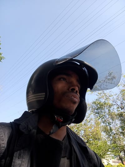 The image shows a close-up, low-angle shot of a person wearing a black helmet with a clear visor. The person's face is partially visible, looking upwards and to the right. They are wearing a black jacket. The background consists of a clear blue sky with several power lines stretching across it. Green foliage from trees is visible in the lower right portion of the image. The scene appears to be outdoors during the daytime. There is no specific information in the image to suggest it is located in Surabaya, Indonesia.