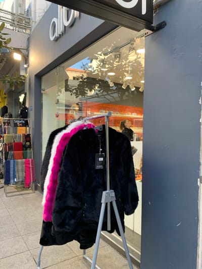 This image captures a street-level view of a storefront in Marousi, Greece, during what appears to be a sunny day, judging by the strong shadows and bright reflections.
In the foreground, a metal clothing rack displays several fluffy or faux fur jackets. The jackets are primarily black, with some featuring prominent white and bright pink trims. Product tags are visible on the black jackets.
Behind the rack is a large shop window, reflecting elements from the street, including tree shadows, parts of buildings, and a clear blue sky. A mannequin head with dark hair can be faintly seen inside the shop, partially obscured by the reflections. Deeper inside the shop, colorful scarves or blankets are neatly arranged on shelves or racks. A small black sign near these items clearly reads "Scarfs 5€", indicating their price in Euros. On the upper left of the shop exterior, a partial white stylized sign, possibly displaying "Olo," is visible against a dark background. The ground visible is a paved sidewalk. The shop's upper facade consists of a dark grey wall.
No direct human activity is depicted, but the scene suggests a commercial retail environment with merchandise displayed for sale. The overall impression is a vibrant, well-lit urban setting.