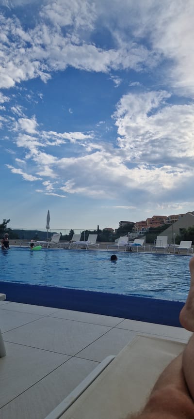 This media file shows a view of a swimming pool area, likely a hotel or resort, on a clear day. The sky is blue with scattered white clouds, suggesting it is daytime, possibly late afternoon given the lighting. The pool is a vibrant blue and appears to be an infinity pool, with a dark blue tiled edge. White lounge chairs with towels are arranged along the side of the pool. In the background, there are buildings with terracotta roofs, nestled on a hillside, which, along with the architecture, suggests a Mediterranean or Adriatic location, consistent with Boreti, Montenegro.

There are several people visible in the pool. One person is swimming, with only their head visible above the water, in the middle of the pool. Further down the pool, a child is playing in the water, possibly with a green inflatable toy. Another person is partially visible at the edge of the pool, near the bottom of the frame. The overall mood seems relaxed and summery, with people enjoying the water and the sunshine. The ground surrounding the pool is paved with large, light-colored tiles. The perspective of the shot is from someone sitting or standing close to the edge of the pool, looking out over the water and the scenery. There is no visible text in the image.