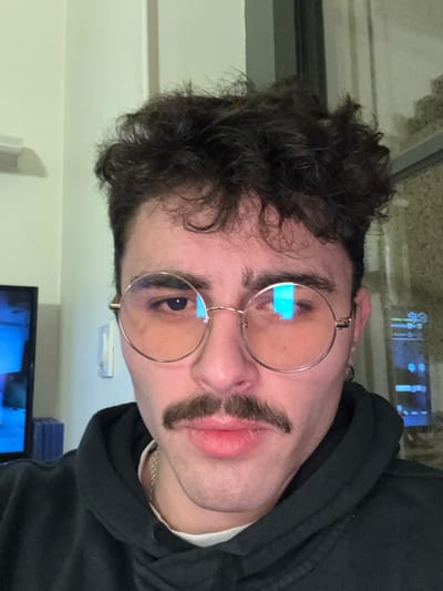 A young man with curly dark hair and round glasses poses. He is wearing a dark hoodie with a white shirt visible underneath. A silver chain necklace is visible on his neck. The man sports a drawn-on, thick, dark mustache. A television screen is partially visible at the bottom of the frame, along with a white wall. The image is a close-up shot, focusing on his face.