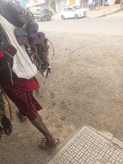 A person, visible from the mid-torso to the feet, stands on the left side of a gravel and dirt ground in Kiamaina ward, Kenya. The individual wears a red and black plaid kilt-like garment and dark brown sandals. Multiple items are suspended from the person's upper body: various leather sandals (including dark brown and purple pairs), small leather pouches, multiple straps, and a light-colored fabric bag. In the lower right foreground, a light-colored concrete utility cover with a geometric pattern is visible on the ground. A paved asphalt road occupies the midground. On the road, a white compact car is positioned slightly right of center, a dark green utility van is visible further left, and a motorcycle is parked on the far right. A row of commercial buildings lines the background across the street. The scene is illuminated by daylight.