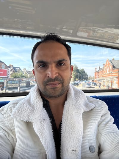 A male individual is depicted in a medium shot, looking directly into the camera with a neutral expression. He has dark hair, dark eyes, and a beard. He is wearing a white corduroy jacket with a white sherpa-style fleece collar and lining, over a dark, open-collar button-up shirt. The image is taken from inside a vehicle, likely a bus, indicated by the blue upholstered seat to the right and reflections on the upper window frame.
Through the front window, an outdoor street scene is visible under a clear, bright sky. To the left, a building with a red sign advertising "BURGERS" is visible, alongside a bus shelter with multiple glass panels and several pedestrians. To the right, across a road, the prominent red-brick Salisbury Railway Station building stands, characterized by its distinctive arched windows, gabled roofs, and chimneys. The road in the mid-ground shows vehicular traffic and parked cars, with a few individuals visible near the station entrance. The scene is located in Salisbury, United Kingdom.