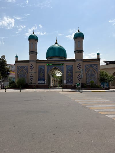 The image depicts the main entrance to the Hazrati Imam Complex (also known as Hast-Imam Square) in Tashkent, Uzbekistan. The architectural style features a central archway flanked by two tall minarets, all adorned with intricate blue and gold tilework. A prominent teal-colored dome sits atop the central structure, with smaller domes crowning the minarets. A digital clock displaying "21:40" is visible above the arch. The foreground shows a paved road with a pedestrian crossing. Several bollards are strategically placed along the walkway in front of the complex. The sky is partly cloudy. People are visible in the background, near the entrance, along with some trees and buildings. The overall scene suggests a public space in a city.