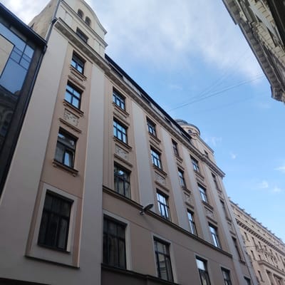 This is a low-angle, wide shot of a beige building facade in Riga, Latvia, under a bright blue sky with scattered clouds. The building features numerous dark-framed windows with reflective glass, and decorative architectural elements above some of the windows. A modern glass-fronted building is partially visible on the left. Overhead, thin wires are strung across the sky, suggesting an urban environment. The building on the right has a more ornate facade with columns and decorative cornices. The perspective of the shot emphasizes the height of the buildings and the expanse of the sky.
