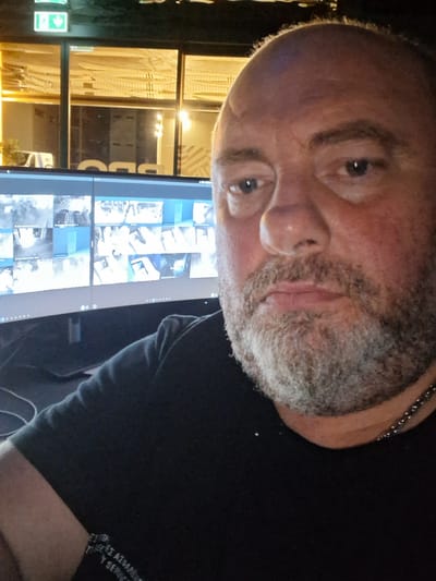 A man with a shaved head and a grey beard is seen from the chest up, looking directly at the camera. He is wearing a black t-shirt and a silver chain necklace. Behind him, a large monitor displays multiple security camera feeds. The background appears to be a room with large windows reflecting lights and possibly objects. The overall lighting suggests an indoor environment, possibly a security control room or office space. Based on the provided information, the location is Mesa Geitonia, Cyprus.
