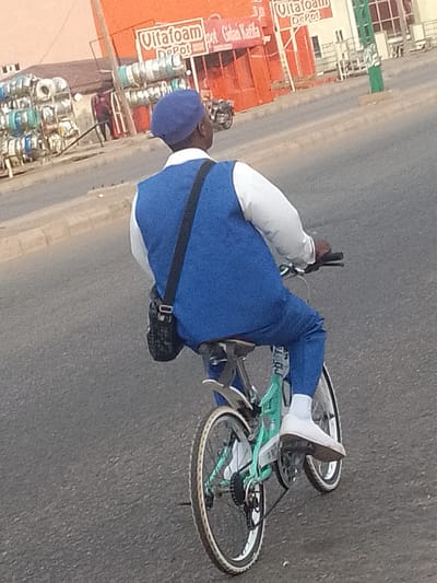A Nigerian man is riding a bicycle on a street in Pantame, Nigeria. He is wearing a blue beret, a white long-sleeved shirt, and blue pants. The bicycle is light green and has a black bag hanging from the handlebars. The man is wearing white socks and white shoes. The street is paved and there are buildings and shops on the side of the road. The buildings appear to be shops selling various goods, including rolls of what look like wire or cable. There is a red building with signage that includes "Ultafoam Depot". The sky is clear and the lighting suggests it is daytime.