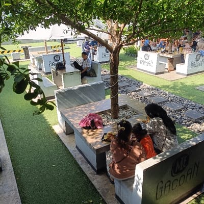 The scene is an outdoor patio area with a large tree in the center. The tree is surrounded by a large concrete platform with white benches and tables. There are several people sitting at the tables. The table nearest the tree is occupied by four people. The three closest to the camera are facing the tree and appear to be eating. One person is seated with their back to the camera. They are wearing a black hijab and a black long-sleeved shirt. The person next to them is wearing a bright yellow shirt. The person furthest away from the camera, seated in a brown dress, is facing the camera and is possibly looking directly at the camera. The other table is occupied by a man in a white collared shirt and grey pants who is sitting at the table and looking down. The scene is located at coordinates -7.002, 110.341, in the city of Kecamatan Ngaliyan, Indonesia. The tables are concrete and have metal frames. The benches are concrete and have a white finish. The patio is paved with grey tiles and there are small rocks scattered about. There is a large tree to the left of the tables, and a large green lawn to the right of the tables. The weather appears to be sunny and there are no clouds in the sky. The sky is blue. The scene is bright and there is plenty of sunlight. The ground is covered with artificial grass. There are several white umbrellas scattered around the patio. Some of the tables have the words "Gacoran" written on them. There are several trash cans on the patio, including one visible directly behind the tree. There are several white metal railings around the patio. The ground is covered in small rocks and the grass is lush and green. There are several plants and flowers in the scene. The scene is tranquil and peaceful.