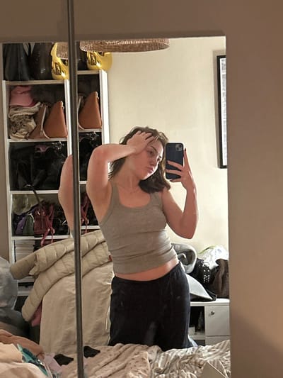 A young woman in a gray tank top and black leggings holds a phone to take a selfie in a mirror. She stands in front of a white shelving unit filled with bags. A framed picture hangs on the wall above the shelving. A wicker basket is visible to the left. The right side of the reflection shows clothing piled on a bed or surface.