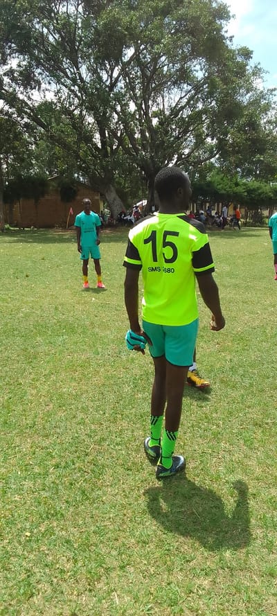 The image shows a soccer field with two men in soccer jerseys.  The man in the foreground, wearing a bright green jersey with the number "15" and the words "SMS 1880" on the back, is looking to the right side of the frame.  He has black and white socks and black cleats.  The man in the background, wearing a teal jersey with the number "4" on it, is standing to the left of the frame. He is looking to the left side of the frame.  He has black socks and brightly colored cleats.  Both men are standing on green grass.  A large tree is visible in the background.  There are many people seated and standing around the field.  There is a brick building in the background and a bicycle is visible in the distance. The coordinates provided (latitude: 0.384, longitude: 34.801) indicate a location in Nairobi, Kenya, not Lubao.
