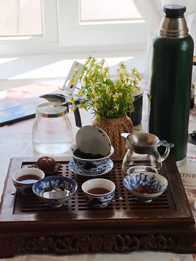 The image depicts a traditional Chinese tea ceremony setup on a wooden tray placed on a table. The tray holds several items, including a glass pitcher filled with water, multiple small porcelain teacups with blue and white Chinese patterns, a teapot with a metal strainer, and a small decorative item resembling a dried fruit. A dark green thermos stands upright to the right of the setup. In the background, a bouquet of small yellow and white flowers in a woven pot is visible, along with a window and white curtains. The scene appears to be indoors, likely in a domestic setting. The presence of a thermos suggests that hot water is available for brewing tea. The overall atmosphere is serene and suggests a moment of quiet contemplation or a social gathering centered around tea. The image does not contain any people or animals, and there are no discernible actions or interactions taking place. The lighting suggests it is daytime, with light coming from the window. The location, Парэчча, Belarus, is provided but there is no visual information within the image to confirm this specific location.