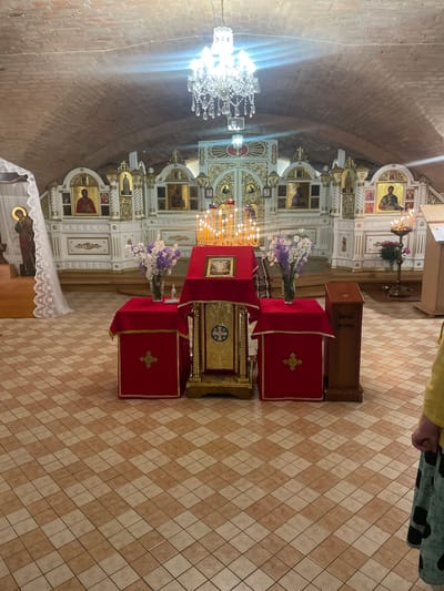 The image depicts the interior of an Eastern Orthodox or Greek Catholic church, located in Terespol, Poland. The scene is illuminated by a large, multi-tiered crystal chandelier hanging centrally from the ceiling, which also casts bright light flares.
The ceiling is a vaulted, exposed red brick structure, showing aged mortar lines. The floor is covered in a checkerboard pattern of light beige and darker brown square tiles.
In the background, a prominent, ornate iconostasis serves as the sanctuary screen. It is predominantly white with extensive gold detailing and features multiple framed icons of saints. From left to right on the iconostasis, several icons are clearly visible, including depictions of figures with halos.
In the mid-ground, a central altar table is draped in rich red fabric. Upon it rests an ornate golden analogion or lectern, which holds an open book. An icon, framed in gold, is also placed on the central table. Numerous lit candles in multiple candelabras are arranged on and around this table, creating a warm glow.
To the immediate left of the central altar table, a smaller table, also draped in red fabric with gold trim, holds a clear glass vase containing a bouquet of white and purple flowers. A small, clear bottle is placed next to the vase. To the far left, partially visible, is a crucifix figure draped with white lace fabric.
To the right of the central altar table, a tall, slender wooden donation box stands on the floor. Inscribed on its front, in Cyrillic script, are the words "ЖЕРТВА НА ХРАМ", which translates to "DONATION FOR THE CHURCH". Further to the right and slightly behind, another candelabra with lit candles is visible.
In the foreground, on the far right, the lower right section of a person is visible. This includes the individual's right hip, a portion of a yellow top, and a green skirt with black polka dots. A partially clenched right hand is also visible at the edge of the frame.