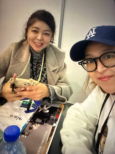 In a medium shot, two individuals are captured in what appears to be an indoor setting. The person on the left, with dark hair pulled back, is wearing a light-colored coat over a patterned top, adorned with a gold chain. They are smiling and holding a small, ornate clutch bag with a gold chain. Their hands are positioned as if showing the bag.

The person on the right, facing the camera, is wearing a blue baseball cap with a white logo, glasses, and a white hooded sweatshirt. They are also smiling and their face is partially obscured by their glasses and the cap.

On a table in front of them, several items are visible, including papers, a blue-capped bottle, and some magazines or booklets. The lighting suggests an indoor environment, possibly a booth or a table at an event, with a neutral-colored wall behind them. The overall impression is a casual, friendly interaction.