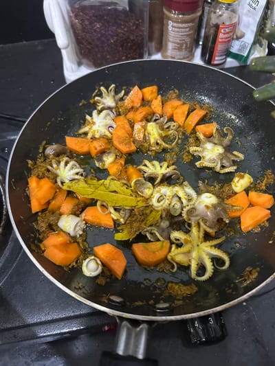 In the city of Sukawati, Indonesia, a black non-stick frying pan sits on a gas stovetop, filled with small octopuses and diced carrots. The octopuses, some with their tentacles curled, are mixed with a dark, savory sauce and appear to be in the process of being cooked. Sliced carrots, vibrant orange, are scattered throughout the pan. A few bay leaves are visible among the ingredients, adding an aromatic touch to the dish. In the background, various spice jars and containers are lined up, suggesting a home kitchen environment. The stovetop's control knobs are visible, with settings marked from 2 to 5. The overall scene depicts a culinary preparation, likely a home-cooked meal, in Sukawati, Indonesia.