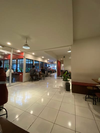 This image depicts the brightly lit interior of a commercial establishment, likely a café or restaurant, located in Pontianak, Indonesia. The view is from a low angle, looking across a reflective white tiled floor towards the rear of the space. The ceiling is light-colored, featuring recessed lighting and two silver pendant lamps.
In the left foreground, the back of a person wearing a dark reddish-brown jacket with a grey stripe on the sleeve is partially visible. Immediately behind them, a glass partition with distinct red framing separates an area where a male in a white shirt is seated at a table. Along the left wall, a series of black-framed windows look into an adjacent interior space. In the mid-left, two males are seated at a small, round black table with black metal stools, facing each other. One wears a dark gray shirt, the other a black shirt.
The walls are primarily light cream. Red accents are present on door frames, a textured vertical panel near the back, and a low, red-upholstered bench seat with a ribbed wooden base extending along the right wall. A large white planter containing green foliage stands in the mid-ground, next to a black waste bin. A square wooden table with two chairs is partially visible on the far right. In the distant background, a counter area with various items is discernible. Artificial lighting evenly illuminates the entire scene.