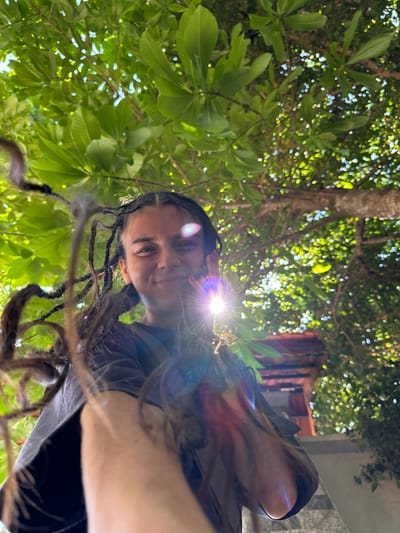A low-angle shot captures a young person with dreadlocks smiling up through lush green foliage. The individual is wearing a dark t-shirt. Sunlight flares brightly near the center of the frame, creating a starburst effect and lens flare that illuminates the scene. The background is a blur of green leaves and dappled sunlight, suggesting an outdoor environment, possibly in a tropical or subtropical location like Mui Ne, Vietnam, known for its beaches and palm trees. Some architectural elements, possibly a tiled roof, are visible in the upper right quadrant. The perspective is as if the viewer is looking up from the ground, with the subject and the canopy of trees forming the primary elements.