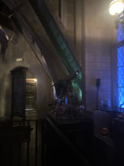 The image depicts an indoor scene, likely a theatrical or themed environment, with a distinctly Gothic or laboratory-like atmosphere. The primary subject is a series of glass display cases, some containing preserved specimens that appear biological, possibly organs or brains, adding a macabre element. One case features what looks like a skeletal hand. These are arranged on dark, aged wooden shelves or countertops.
The setting is dimly lit, with the light source appearing to be an ornate, possibly vintage, lamp fixture with a glowing bulb in the upper left of the frame. Blue light emanates from a grid-like window or ceiling feature at the top. The architecture is characterized by rough-hewn stone or concrete walls with visible joint lines, and a sweeping staircase or ramp with a metallic railing. Some areas of the scene are in deep shadow, obscuring finer details.
There are no people clearly visible. The overall mood is mysterious and slightly unsettling due to the subject matter of the displays. The time of day and weather are not discernible. The location cues, such as the architectural style and the general presentation, suggest a specialized venue rather than a typical public space. There is no discernible text in the image.