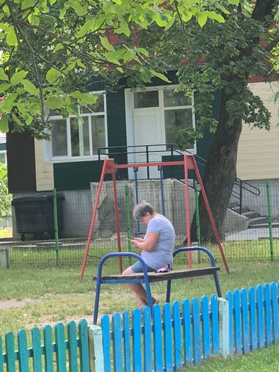 A person is bent over a mobile device while sitting on a blue and green wooden bench. They are wearing a light blue t-shirt and denim shorts. The bench is part of a playground structure with blue metal supports and red metal bars. The playground is set in a grassy area with a fence made of blue and green wooden planks. In the background, there is a light-colored building with green and dark siding, and several windows. A large tree with green leaves is to the left of the frame, with its trunk resting on the building. The scene appears to be outdoors during the day, with natural lighting. The overall setting suggests a residential area or a park. There is no visible text in the image.