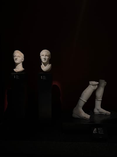 This image depicts a dimly lit exhibition space, likely within a museum in Istanbul, Turkey, showcasing ancient sculptures. On the left, two marble busts are displayed on dark, rectangular pedestals. The bust on the left features a female figure with her hair styled in an intricate updo. The bust on the right portrays a male figure with short, curly hair. Both busts are well-lit, highlighting their classical features.
To the right of the pedestals, a separate display features a pair of disembodied marble legs and feet, also resting on a dark, tiered platform. The legs are adorned with decorative bands around the ankles and calves, and the feet are intricately carved, showing sandals or similar footwear. A small plaque with text and an illustration of the legs is visible on the platform in front of this sculpture. The background is a deep, dark red, creating a dramatic contrast with the pale marble of the sculptures. The overall lighting suggests a controlled exhibition environment designed to emphasize the artifacts.
