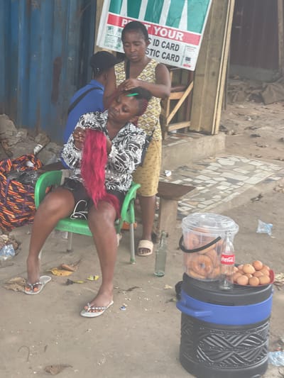 Two women are seen outside, one braiding the other's hair, which is partly red. The braider wears a patterned yellow shirt and the seated woman wears a black and white shirt. A man in a blue shirt leans against the seated woman. Nearby, a cooler with eggs and doughnuts sits on the ground. A sign reads "Print Your ID Card Plastic Card". The scene takes place in a street setting with scattered debris.