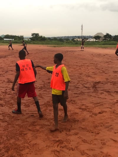 The image depicts a dirt field in Nsugbe, Nigeria, with several people wearing bright orange vests. Two individuals are prominently featured, with one person’s hand touching the back of another’s vest. There are additional people in the background also wearing similar orange vests. Visible in the distance are buildings and structures, one of which appears to be a communications tower. The sky is overcast.