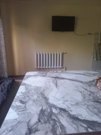 The image depicts an indoor scene, likely a room, with a large marble-patterned table dominating the foreground. The table's surface has a grey and white veined design. In the background, a white radiator is mounted on a pale yellow wall. Above the radiator, a black flat-screen television is affixed to the wall, with a black cord running to a power outlet. To the left of the radiator, a curtain with a textured pattern hangs. On the right side of the image, partially visible on the wall, are two folded towels hanging on what appears to be a towel rack or hooks. A dark blue blanket or piece of fabric is visible on a piece of furniture to the left of the table. The lighting in the room is dim, casting shadows and creating a muted atmosphere. There are no people visible in the image. The scene appears to be a domestic interior setting.