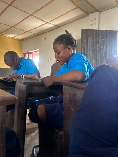 The image, taken in Ikorodu, Nigeria, captures an indoor scene within what appears to be a classroom or study area, illuminated by bright natural light suggesting daytime.
Three individuals, likely students, are visible, all wearing similar blue short-sleeved tops that resemble school uniforms. In the foreground, a young woman with braided hair is leaning forward onto a sturdy, worn wooden desk. She has a focused yet soft expression, possibly a faint smile, and wears a unique earring shaped like a question mark.
Behind her, positioned to the left, is another young person, a male, also in a blue uniform and leaning over a similar wooden desk. He wears a pink wristband adorned with a black and red logo or symbol. Both individuals appear engrossed in an activity, likely studying or writing at their desks. Part of a third person, dressed in dark blue, is visible on the far right.
The room features light-colored walls, one of which has a grid-like pattern created by wooden strips over light panels. A red-framed window allows ample daylight into the space. The ceiling is light-colored, with a visible light bulb fixture and a few exposed bolts or hooks near a corner. The floor is tiled. Faint, possibly numeric, markings like "4" can be seen on the surface of one of the wooden desks. The overall atmosphere suggests a dedicated learning environment.