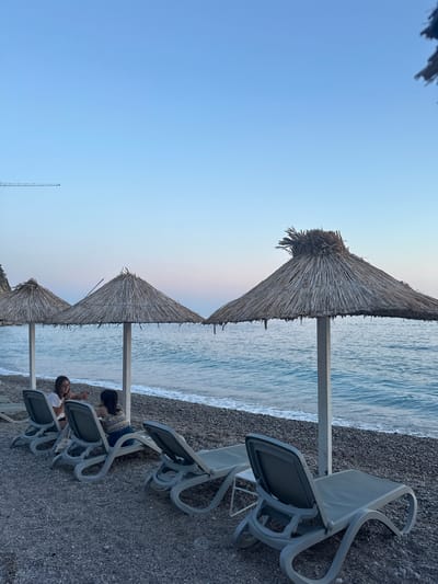 The image depicts a serene beach scene in Mirište, Montenegro, at dusk. In the foreground, several grey plastic lounge chairs are arranged on a pebble beach, some unoccupied and others occupied by two individuals. The first person, facing left, is wearing a white t-shirt and dark-rimmed glasses. The second person, facing right, is wearing a patterned top and dark pants. They appear to be engaged in conversation. Behind the lounge chairs are three thatched umbrellas, providing shade. The calm, blue sea with gentle waves is visible in the background, stretching towards the horizon under a clear, pale blue sky that transitions to soft pink hues near the horizon. A faint, distant crane is visible in the upper left corner of the sky. The overall atmosphere is tranquil and relaxing.