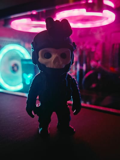A dark, low-angle, close-up shot captures a stylized, skull-faced figurine with a tactical helmet and suit. The figurine stands on a dark, textured surface. In the background, a computer case is visible, illuminated by vibrant neon pink and cyan lights. The pink lights are from fans at the top of the case, and the cyan light is from a fan on the left side, which has a visible "CY" logo. The scene is dimly lit, with the neon lights casting colorful reflections on the figurine and the surrounding environment. The overall atmosphere is moody and futuristic. The image does not contain any information about the city of Vladivostok, Russia.