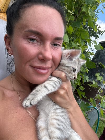 The image shows a close-up selfie of a woman holding a small, grey tabby kitten. The woman has dark hair pulled back and is wearing a gold necklace. She has earrings in her left ear, one with a black clover design. The kitten is held against her chest, with its head turned to the right, looking away from the camera. The background is a garden with green foliage and a blue sky visible. The woman's skin appears to be sun-kissed, and there are visible pores and fine lines on her face. The kitten's fur is soft and striped, and its eyes are open. The overall impression is of a casual, outdoor setting.