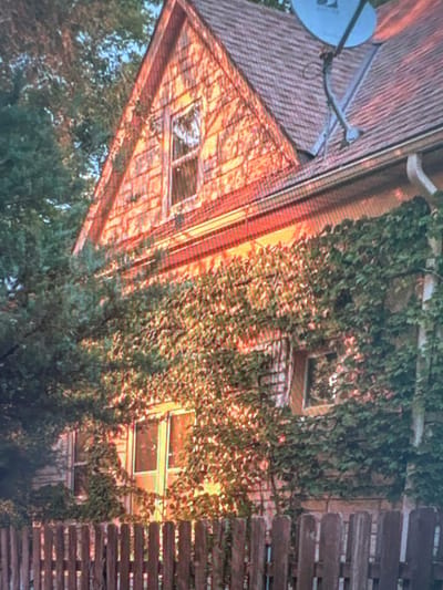 The image shows the side of a two-story house with wooden siding and a sloped roof covered in shingles. A satellite dish is mounted on the roof. The house is partially covered by ivy and other foliage, and a wooden fence is visible in the foreground. The lighting suggests it may be late afternoon or early morning, with warm sunlight illuminating the side of the house. There are no people or animals visible in the image. The location is identified as Kuvshinovo, Russia.