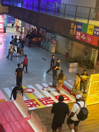 This image captures a bustling indoor pedestrian area in Liangjiang, China, possibly a shopping mall or entertainment complex, viewed from an elevated perspective. In the foreground, two men are walking away from the camera. One is wearing a black t-shirt and shorts with a black backpack, while the other wears a white t-shirt, black shorts, and a white baseball cap with a black backpack. A bright yellow neon sign is visible to the right.
Further into the scene, a group of people are gathered near a crosswalk painted on the floor, which features cartoonish figures and red and yellow lettering. Several individuals are walking across this crosswalk. To the left, a red structure with raised wooden sections is prominent. In the background, various shops and entertainment booths are visible, including arcade machines and merchandise displays. Another group of people are interacting near a set of stairs and a railing on a higher level. A person is lying on a mat in the far background, possibly receiving a massage. The environment appears to be an indoor public space with a polished concrete floor and overhead lighting.