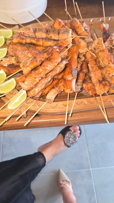 The image shows a close-up of a wooden platter filled with numerous salmon skewers, seasoned and grilled. Several lime wedges are placed on the side of the platter. In the foreground, a person's feet are visible, one wearing a black sandal embellished with colorful rhinestones and the other a nude pointed-toe flat with a gold ankle bracelet. The background suggests an indoor setting with a tiled floor. The scene appears to be a casual meal or gathering, possibly at a restaurant or home. The image does not provide any specific information about the location being Trikomo, Northern Cyprus / Cyprus.