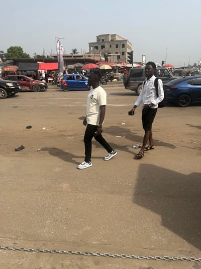 Two young men stand on a street in an urban African setting. One wears a white button-down shirt, black shorts, and sandals, holding a small object. The other is dressed in a white short-sleeved shirt, black pants, and white sneakers. The background shows a bustling scene with buildings under construction, various vehicles (cars, a blue auto-rickshaw), market stalls with red umbrellas, and other individuals. A heavy chain is visible on the right side of the image. The sky is hazy.