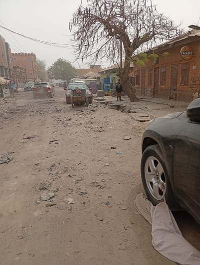 The image depicts an unpaved, dusty street scene in Sabon Gari West, Nigeria, under an overcast or hazy sky. The foreground features a dirt road with scattered debris, gravel, and uneven surfaces. To the right, the front portion of a dark-colored SUV is visible, with a light-colored cloth draped near its front wheel on the ground.

Mid-ground shows a series of vehicles and individuals. A dark-colored sedan is positioned centrally on the road, with a two-wheeled cart attached to its rear. Behind it, a light-colored SUV travels away from the viewer, generating a small cloud of dust. Further down the street, multiple other vehicles are visible.

On the right side of the street, a prominent leafless tree with numerous dark, elongated seed pods stands on a raised dirt embankment. Next to the tree is a one-story building with a rust-colored or earthen facade, featuring multiple brown wooden shuttered windows and doors. A circular "Gulder" sign is affixed to the building, along with text including "PAN AFRICAN HOTEL" and a sign partially reading "VEHICLES WERE...". An individual walks on a rough concrete sidewalk adjacent to this building. Stacked blue and green crates are visible further down the sidewalk near a parked motorcycle.

On the left side of the street, multiple multi-story buildings are present. One building features a reddish-brown exterior with distinctive architectural elements resembling turrets along its roofline. At least two individuals are visible standing near these buildings. Overhead utility wires crisscross across the scene. The overall environment suggests a commercial or residential district with an unmaintained road infrastructure.