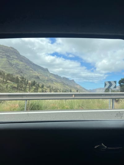The image is a view from inside a vehicle, likely a car, looking out the window. In the background is a landscape of green mountains under a cloudy sky with patches of blue. A road with a guardrail is visible in the foreground. A road sign with black and white chevron arrows pointing to the left is next to the guardrail. The image appears to be taken while traveling along a road in a mountainous area. Based on the location of Mogán, Spain, and the landscape features, it is likely that the image was taken somewhere along the winding mountain roads in the municipality of Mogán, Gran Canaria, Spain. There are no people visible.