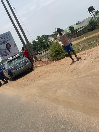 The image, rotated 90 degrees clockwise, captures an outdoor scene, likely a street or unpaved road in Galadimawa, Nigeria, under a clear, sunny sky during the daytime. The ground consists of a light brown dirt path bordering a grey, possibly paved, road surface.
In the mid-ground, a man wearing a light-colored long-sleeve top, blue shorts, and dark flip-flops stands on the dirt path, facing away from the camera, his shadow clearly visible. Adjacent to him, on the paved section, a light blue/teal sedan car is parked. The car's license plate reads "ABC-97580", and "DUNAM'S" is visible on its trunk.
Two other individuals are near the car: one in a red shirt is partially visible near the rear, appearing to lean into the car, and another, in blue clothing, is partially seen near the front, possibly squatting or bending. Power lines and utility poles diagonally cross the lower left portion of the frame.
A large billboard stands prominently near the car and poles, displaying real estate information: "AN SHELTER", "ATE LOKOGOMA", "BEDROOM DUPLEX", "#180M", a phone number "0803 312 3216", and "EZEKIEL PROPERTIES MANAGEMENT CO".
In the background, several buildings with light-colored, possibly metal, roofs are visible, some featuring solar panels. A tall metal structure, potentially a water tower or communication mast with two tanks, rises above the buildings, surrounded by green trees and bushes. Strong shadows indicate bright sunshine.