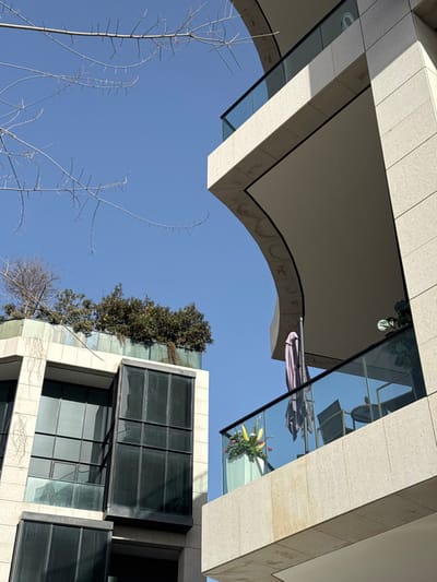 Captured from below, an architectural scene features beige, tiled buildings against a clear blue sky. A curved archway frames the upper portion, while a structure with dark-tinted windows stands diagonally. The windows reflect the surrounding architecture. Sparse branches reach into the sky from the lower left. A glass-enclosed space with plants and outdoor furniture is visible, further adding to the complex interplay of lines and reflections.