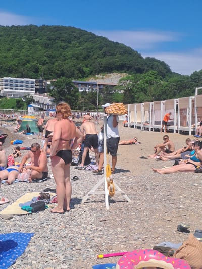 The image depicts a crowded beach scene in Dzhubga, Russia, under a clear blue sky with scattered clouds. The beach is covered with pebbles, and numerous people are engaged in various beach activities. In the foreground, a woman with red hair wearing a black bikini stands with her hands on her hips, facing away from the camera. Nearby, a man wearing sunglasses and black swim trunks sits on a towel, attending to a plastic bag. Several other individuals are sunbathing and relaxing on towels and mats scattered across the beach.
In the midground, a vendor is carrying a tray of donuts on his head. Further back, beach umbrellas and temporary structures provide shade. The sea is visible in the distance, with a gentle slope leading down to the water. Behind the beach, a lush green hillside covered in trees rises prominently, with several buildings, including a multi-story hotel, visible on its slope. The overall atmosphere suggests a busy summer day at a popular tourist destination.
