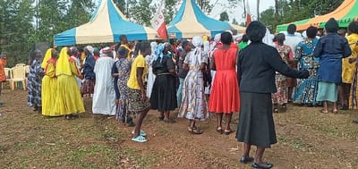 A large gathering of people, primarily women, are present in an outdoor setting, likely a community event, located in Lwandeti, Kenya, at the coordinates provided. The scene unfolds beneath the shade of a large tent with a blue and white striped canopy. The people are dressed in colorful clothing, many in bright yellow and red dresses. Some are wearing traditional African patterns. A few men are also present, standing in the background and participating in the gathering. The atmosphere is lively and celebratory. The women appear to be engaged in social interactions, possibly dancing or singing, while the men are observing. The ground is covered in a thick layer of grass, typical of an outdoor setting. The area is surrounded by trees and greenery, creating a natural and vibrant environment. It is a joyous occasion with a strong sense of community engagement.