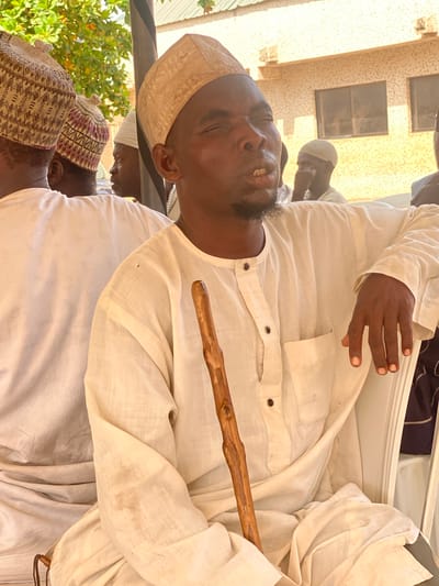 An elderly Black man with a light-skinned complexion and a goatee sits in the foreground, holding a wooden walking stick. He wears a cream-colored traditional outfit with a matching cap. His eyes are closed, and his mouth is slightly open, as if speaking or singing. The man is seated on a white plastic chair, and his left arm rests on his lap, with his hand visible. In the background, several other men are partially visible, also wearing traditional attire and caps. The setting appears to be outdoors, with a building and foliage in the background. The building is light yellow with windows. The overall scene suggests a community gathering or event in Maiburiji, Nigeria.