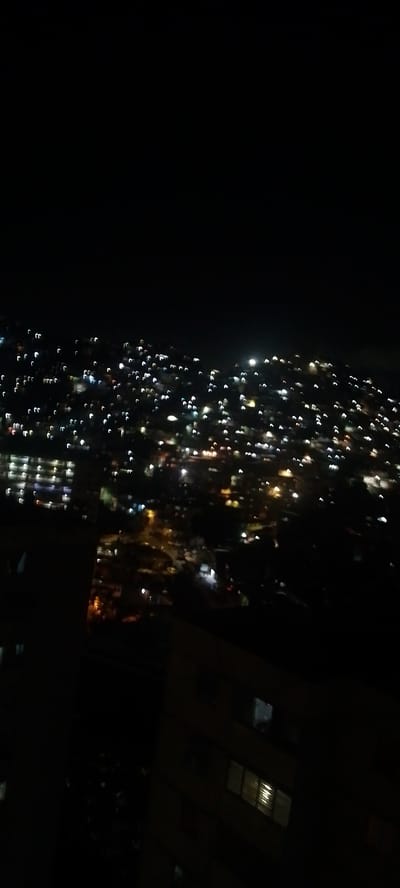 The image is a nighttime, blurry aerial view of a city, likely Caracas, Venezuela, characterized by the bokeh effect of numerous lights against a dark sky. The city appears to be built on hills, with lights scattered across the slopes, suggesting a densely populated urban area. The lights are mostly white and yellow, with some occasional blue and red hues, creating a warm, diffused glow. In the foreground, there are dark, silhouetted buildings, one of which has illuminated windows, hinting at residential or commercial structures. The overall impression is of a vast, sprawling metropolis at night, with its lights creating a distinct pattern against the darkness.