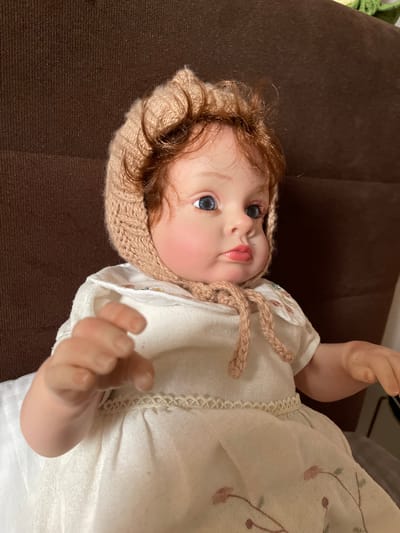A realistic baby doll with blue eyes, rosy cheeks, and auburn hair is pictured indoors. The doll wears a knitted bonnet, a white dress with floral embroidery, and is seated against a brown textured background, likely a couch or chair. The doll's left hand is positioned in the foreground, appearing to reach forward. This image was taken in Zelenogradsk, Russia.