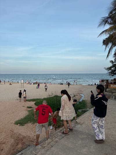 The image depicts a beach scene in Phan Thiết, Vietnam, likely during the late afternoon or early evening. The sky is a pale blue with light clouds, and the sea is calm with gentle waves. Numerous people are visible on the sandy beach and in the water, engaged in various activities such as swimming, sunbathing, and walking.
In the foreground, a group of people are walking up a concrete path. One individual in a red t-shirt with the number "5" and the text "TH TRUE MILK" is particularly prominent. They are wearing light-colored shorts and flip-flops. Beside them, a young boy in red shorts is walking down the path. Further up the path, a woman in a white lace dress and a young person in a black hoodie and patterned pants are also present. The woman is holding a mobile phone.
The beach itself is wide and sandy, with some patches of green vegetation. Several beach umbrellas and chairs are scattered across the sand, indicating a popular recreational area. In the distance, the coastline is visible with some buildings, and palm trees line the edge of the beach. A wooden structure with signage that includes "CATV" and "B&B" is situated on the right side of the image. The overall atmosphere appears relaxed and leisurely.