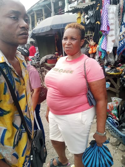 The image depicts an outdoor market scene in Ura Nta Umuarandu, Nigeria. In the immediate foreground on the left, an adult male with dark skin is partially visible, wearing a yellow shirt with blue and brown patterns, and a black strap across his chest. He is looking slightly to his right. To his right, in the mid-ground, stands an adult female with dark skin and short, light-colored hair. She is wearing a pink t-shirt with a green "VEROSCE" logo on the chest, exhibiting sweat stains, white shorts, and dark open-toed sandals. A small dark shoulder bag with a chain strap is worn across her body, and she holds a blue and black striped plastic bag in her left hand. A silver-colored watch is on her left wrist. Her facial expression is neutral, and she is looking forward. Behind the foreground figures, other individuals are partially visible, including one in a pink top and dark trousers with a white stripe, wearing a black cap, and another in a red shirt further in the background. The background is composed of multiple market stalls with various goods displayed, including shoes, socks (some with zigzag patterns or mustaches), and clothing, beneath awnings and umbrellas. A blue and white "Chelsea" football club flag is visible among the merchandise. The ground is an unpaved, uneven surface.